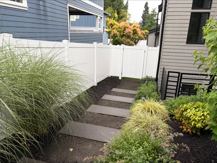 White privacy fencing and a gate at the end of a landscaped path between two houses.
