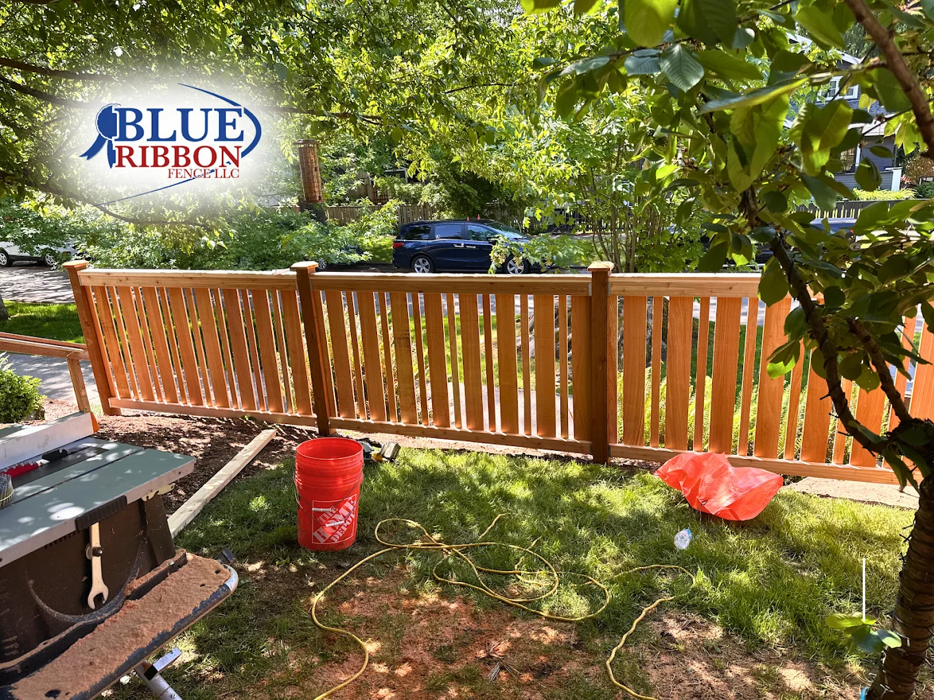 Wood fence with open vertical slats beneath a leafy tree, photographed during installation with the Blue Ribbon logo.